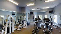 This fitness center offers a comprehensive workout environment designed to accommodate a range of exercise needs. The space is finished with neutral gray walls and carpeted flooring, creating a clean and functional atmosphere. Large mirrors line one wall, enhancing the sense of space while allowing users to monitor their form during workouts. The room is illuminated by a combination of recessed fluorescent lighting and ceiling fans, ensuring consistent airflow and visibility throughout. Natural light filters in through multiple windows along one wall, providing pleasant views of surrounding greenery. The strength-training area features a multi-station cable machine and a dedicated free weights rack stocked with a range of dumbbells, along with adjustable benches for versatile use. The cardio section is well-appointed with treadmills, an elliptical trainer, and a stationary bike, offering varied options for cardiovascular exercise. Wall-mounted televisions are positioned for easy viewing from both the cardio and strength areas. Ceiling fans distributed throughout the space contribute to comfortable air circulation during workouts.
