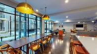 This single-floor amenity space is thoughtfully designed to offer a variety of social and recreational environments within a cohesive, contemporary aesthetic. The club room serves as the heart of the space, featuring plush sectional sofas, colorful accent chairs in teal, blue, and burnt orange, and a wall-mounted television flanked by open wood shelving. A striking gallery wall covered in eclectic framed artwork adds a personalized, creative touch to an intimate lounge nook nearby. Floor-to-ceiling black-framed windows flood the lounge and dining areas with natural light and provide views of the surrounding exterior courtyard. The dining area is anchored by a long communal table surrounded by wooden chairs with patterned cushions, illuminated by bold, oversized brass pendant lights that add warmth and visual drama. Adjacent to the dining area, a bar features a vivid teal subway-tiled accent wall, a white countertop with geometric-patterned cabinetry, bar seating, and open wood shelving. The fitness center is generously equipped with cardio machines, free weights, and cable machines, accented by a vibrant abstract mural and large windows overlooking the courtyard. Polished concrete and dark hardwood floors unify the space throughout.