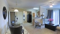 This single-floor residence offers an open and cohesive layout that seamlessly connects the living, dining, and kitchen areas into one flowing, functional space. The living and dining area is finished with plush wall-to-wall carpeting and features elegant crown molding along the ceilings, lending a polished, refined character to the interior. A pendant light fixture anchors the dining area, where a sleek glass-topped table is paired with classic leather chairs, creating a sophisticated yet comfortable setting. Adjacent to the dining space, a houndstooth accent chair and mirrored console table add a touch of eclectic style to the living area. The kitchen is defined by a raised breakfast bar that provides a natural transition from the open living space, and is equipped with full-size appliances including a black refrigerator. Warm neutral wall tones carry throughout the unit, complemented by consistent trim and millwork details. A hallway visible from the main living area leads to the bedroom, which features carpeting and appears bright and private. The overall ambiance is warm, inviting, and thoughtfully arranged, balancing everyday practicality with stylish residential design.