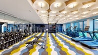 This fitness center is a visually dynamic and fully equipped space designed to support a wide range of workout routines. The most distinctive architectural feature is the ceiling, which is adorned with an eye-catching arrangement of large, overlapping circular forms finished in white and gold, embedded with integrated spotlights that cast warm, even illumination throughout the room. This sculptural canopy lends the space a modern, upscale character.

The flooring makes an equally bold design statement, featuring a large-scale chevron and hexagon pattern in yellow, blue, gray, and white — adding energy and visual interest underfoot. Deep navy accent walls and floor-to-ceiling windows line one side of the space, allowing ample natural light and views of the surrounding exterior, contributing to an open and motivating atmosphere.

The fitness center is divided into functional zones. The strength training area includes Matrix-branded benches, a dedicated training zone with a multi-functional rig, medicine balls, kettlebells, a full dumbbell rack, and cable machines, all positioned alongside a mirrored wall. The cardio zone features a row of treadmills, ellipticals, and stair climbers oriented toward the exterior windows. A heavy bag and TRX suspension system round out the space's boxing and functional fitness offerings.