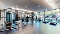 This contemporary fitness center is thoughtfully designed to support a wide range of workout routines in a sleek, modern environment. The main exercise floor features stylish herringbone-patterned flooring and is lined with floor-to-ceiling windows along one wall, flooding the space with natural light and offering views of lush greenery outside. Recessed lighting and ceiling fans maintain a bright, comfortable atmosphere throughout.

The main workout area is comprehensively equipped with cardio machines including treadmills, ellipticals, and a stationary bike, alongside an extensive selection of strength training equipment such as a cable machine, weight racks, medicine balls, kettlebells, and a punching bag. Large mirrors line one side of the room, enhancing the sense of openness. A dedicated Matrix cable system provides a versatile option for functional training.

A separate spin room offers a focused cycling experience, featuring multiple stationary bikes and bold geometric wall art in black and gold that adds an energetic visual element to the space. Adjacent to the main floor, an additional open area houses strength machines and is accented by striking abstract artwork, creating an inviting and motivating environment. An accessible restroom and convenient water station round out the facility's amenities, ensuring a complete and well-appointed fitness experience.