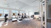 Located at 200 Big Wds Dr in Poquoson, Virginia, this single-floor fitness center offers a bright and inviting space for residents to pursue their wellness goals. The room features soaring vaulted ceilings that lend an open, airy feel, complemented by multiple ceiling fans that ensure comfortable airflow throughout workout sessions. Large, floor-to-ceiling windows line the walls, flooding the space with natural light and providing pleasant outdoor views of the surrounding community.

The fitness center is comprehensively equipped to support a full range of exercise routines. Cardio equipment includes treadmills, elliptical machines, and a stationary cycling bike, each positioned to take advantage of the natural light streaming in from the windows. A dedicated strength training area features a multi-functional cable machine system, a dumbbell rack with a tiered selection of free weights, an adjustable workout bench, and additional resistance machines. A wall-mounted flat-screen television is positioned near the free weights area for entertainment during workouts. Full-length mirrors are installed along one wall to assist with form and technique.

The flooring consists of durable, multi-toned carpet tiles, and the clean white walls contribute to the modern, minimalist aesthetic. Glass-panel doors provide easy access in and out of the facility, enhancing the welcoming atmosphere of this well-appointed community fitness center.