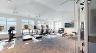 Located at 200 Big Wds Dr in Poquoson, Virginia, this single-floor fitness center offers a bright and inviting space for residents to pursue their wellness goals. The room features soaring vaulted ceilings that lend an open, airy feel, complemented by multiple ceiling fans that ensure comfortable airflow throughout workout sessions. Large, floor-to-ceiling windows line the walls, flooding the space with natural light and providing pleasant outdoor views of the surrounding community.

The fitness center is comprehensively equipped to support a full range of exercise routines. Cardio equipment includes treadmills, elliptical machines, and a stationary cycling bike, each positioned to take advantage of the natural light streaming in from the windows. A dedicated strength training area features a multi-functional cable machine system, a dumbbell rack with a tiered selection of free weights, an adjustable workout bench, and additional resistance machines. A wall-mounted flat-screen television is positioned near the free weights area for entertainment during workouts. Full-length mirrors are installed along one wall to assist with form and technique.

The flooring consists of durable, multi-toned carpet tiles, and the clean white walls contribute to the modern, minimalist aesthetic. Glass-panel doors provide easy access in and out of the facility, enhancing the welcoming atmosphere of this well-appointed community fitness center.