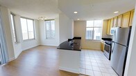 This single-floor space showcases an open-concept layout that seamlessly integrates the kitchen and living areas. The kitchen is anchored by a peninsula with striking dark granite countertops and a stainless steel sink, providing both functional workspace and a natural dividing point between the two zones. Light wood cabinetry adds warmth to the kitchen, which also features stainless steel appliances and large-format white tile flooring.

The living area transitions to smooth hardwood-style flooring, creating a visual distinction between the two spaces while maintaining a cohesive overall aesthetic. Neutral white walls throughout keep the interior bright and versatile. Multiple large windows on opposing walls flood the space with natural light and offer views of the surrounding urban cityscape, lending the unit a notably airy and open atmosphere.

Recessed lighting supplements the natural light, while a track lighting fixture in the living area provides targeted illumination. The open layout and multiple windows contribute to a sense of spaciousness, making this a well-lit, functional urban space suitable for everyday living.