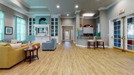 Located at 2042 Arrowgrass Dr in Wesley Chapel, FL, this well-appointed single-story space offers a thoughtfully designed open-concept layout with a warm, contemporary aesthetic. The interior is characterized by warm wood-look flooring, crown molding, recessed lighting, and a cohesive neutral color palette accented by teal and turquoise tones throughout.

The expansive main living area features a built-in entertainment wall with display shelving, flanked by a wet bar peninsula with bar seating — ideal for entertaining. This open space flows seamlessly into additional seating areas, including a bright sitting room lined with floor-to-ceiling windows dressed in sheer teal curtains that flood the room with natural light.

A dedicated dining area accommodates a glass-top round table set beneath large windows, creating an airy and inviting atmosphere. The fully equipped kitchen includes white cabinetry, granite countertops, stainless steel appliances, and a window above the sink offering views of the outdoors. Stone tile flooring anchors the kitchen's clean, functional design.

A rotunda-style entry vestibule with tall windows adds architectural distinction to the space. Throughout, the design balances openness with defined functional zones, making this a versatile and visually cohesive property suited for a variety of uses.