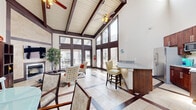 This well-appointed single-floor space makes a striking first impression with its dramatic vaulted ceilings accented by rich dark wood exposed beams and white shiplap paneling. Dual ceiling fans circulate throughout the open main area, which seamlessly integrates a living room, dining area, and kitchen into one cohesive, light-filled environment. Floor-to-ceiling windows and glass-paneled French doors line the exterior wall, flooding the space with natural light and offering views of the outdoor surroundings. The living area centers around a sleek, tile-framed fireplace with a wall-mounted television above, complemented by an elegant patterned tile floor featuring dark wood-toned inlaid accents. The kitchen is efficiently designed with rich espresso cabinetry, granite countertops, stainless steel appliances, and a raised breakfast bar with bar seating — ideal for casual dining or socializing. A separate, glass-enclosed office provides a quiet, functional workspace complete with a ceiling fan, tile flooring, and ample room for a workstation and seating. Throughout the space, soaring wall heights and an open layout create a sense of expansiveness well beyond the footprint, making this a versatile and inviting environment suited for both productive work and comfortable gathering.
