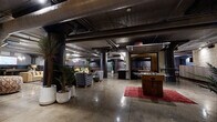 This expansive single-floor space offers a striking industrial aesthetic defined by polished concrete floors, bold dark structural columns, and fully exposed ductwork and mechanical systems running across the ceiling. The open layout encompasses a generous footprint designed to accommodate multiple distinct activity and social zones simultaneously.

A central lounge area features an eclectic mix of upholstered seating, patterned armchairs, and decorative rugs, anchored by warm pendant lighting and lush tropical potted plants that soften the raw industrial character. A foosball table and shuffleboard table provide recreational focal points, while a dedicated bowling lane — complete with mounted flat-screen displays — occupies its own defined zone, accompanied by a comfortable seating nook.

The space also features intimate booth-style seating alcoves set against exposed brick walls, offering a more private gathering option. A bar and counter area serves as a central service hub. Curtained dividers along one wall allow for flexible spatial configurations, and track and pendant lighting throughout creates warmth against the dark, utilitarian palette. The overall ambiance balances raw, warehouse-inspired architecture with curated, lounge-ready furnishings — making it well-suited for social events, co-working, and community gatherings.