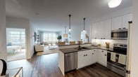 Located at Greybarn Amityville in Amityville, NY, this single-floor residence showcases a thoughtfully designed open-concept layout that seamlessly connects the kitchen, dining, and living areas. The kitchen is outfitted with crisp white shaker cabinetry, dark countertops, classic subway tile backsplash, and a full suite of stainless steel appliances, including a refrigerator, range, microwave, and dishwasher. A peninsula with pendant lighting creates a natural transition into the living space, which is bathed in natural light from large windows overlooking the outdoors. Rich hardwood-style flooring runs throughout the main living areas, adding warmth to the clean, neutral palette. The spacious living room provides a comfortable and airy atmosphere, ideal for relaxing or entertaining. The primary bedroom features plush carpeting, generous proportions, and direct access to a well-appointed en-suite bathroom. The bathroom is finished with white subway tile surrounding the soaking tub and shower, a classic mosaic tile floor, a wide vanity with dark countertops, and a large mirror. A second bedroom with a walk-in closet offers additional flexibility. The entry foyer provides a clean, welcoming arrival with ample cabinet storage nearby. Throughout the residence, the cohesive design balances modern functionality with timeless style.
