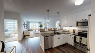 Located at Greybarn Amityville in Amityville, NY, this single-floor residence showcases a thoughtfully designed open-concept layout that seamlessly connects the kitchen, dining, and living areas. The kitchen is outfitted with crisp white shaker cabinetry, dark countertops, classic subway tile backsplash, and a full suite of stainless steel appliances, including a refrigerator, range, microwave, and dishwasher. A peninsula with pendant lighting creates a natural transition into the living space, which is bathed in natural light from large windows overlooking the outdoors. Rich hardwood-style flooring runs throughout the main living areas, adding warmth to the clean, neutral palette. The spacious living room provides a comfortable and airy atmosphere, ideal for relaxing or entertaining. The primary bedroom features plush carpeting, generous proportions, and direct access to a well-appointed en-suite bathroom. The bathroom is finished with white subway tile surrounding the soaking tub and shower, a classic mosaic tile floor, a wide vanity with dark countertops, and a large mirror. A second bedroom with a walk-in closet offers additional flexibility. The entry foyer provides a clean, welcoming arrival with ample cabinet storage nearby. Throughout the residence, the cohesive design balances modern functionality with timeless style.