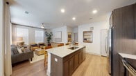 This well-appointed single-floor residence offers a thoughtfully designed open-concept layout that seamlessly connects the kitchen, dining, and living areas. The kitchen serves as the centerpiece, featuring a generously sized granite island with an undermount sink and bar seating, rich dark-brown cabinetry, stainless steel appliances including a refrigerator, range, and microwave, and under-cabinet lighting. Light wood-style flooring flows throughout the main living areas, complementing the bright, neutral color palette.

The living room is bathed in natural light through multiple large windows dressed with blinds and floor-length curtains, and is finished with a ceiling fan. The space offers a comfortable and open atmosphere ideal for relaxing or entertaining.

The primary bedroom features a warm, accent-painted headboard wall, carpeted flooring, and ample natural light. A private en-suite bathroom includes a granite-topped vanity with dark cabinetry, a tub-shower combination with tile surround, and direct access to a spacious walk-in closet with built-in shelving. A second bedroom offers similar comfort with its own window and carpeted floors.

A dedicated laundry room houses a full-size top-load washer and dryer with overhead shelving. The residence combines functional design with modern finishes throughout.