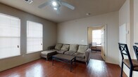 This single-story unit is organized around a practical, efficient layout that connects the main living spaces seamlessly. The living room serves as the central gathering area, featuring warm hardwood-style flooring, neutral-toned walls, and a ceiling fan with integrated lighting. Multiple windows fitted with horizontal blinds allow natural light to filter in, creating a comfortable atmosphere. A door from the living room provides direct access to what appears to be a small outdoor area or adjacent space.

The kitchen is positioned adjacent to the living area and is defined by a raised breakfast bar with bar stool seating, making it a functional space for casual dining. Medium-toned wood cabinetry lines the walls, complemented by dark countertops and black appliances, including a range and an overhead microwave. A door off the kitchen leads to a utility or storage area.

The bedroom is a quiet, carpeted room with neutral walls, offering a straightforward and practical space for rest. The bathroom includes a dark-vanity with a sink, a mirror, textured walls, and wood-style flooring consistent with the rest of the unit. Overall, the space presents a cohesive, no-frills design with functional finishes throughout.