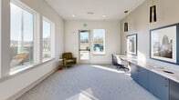 Located at 225 Rhyne Rd in Charlotte, NC, this single-story space is designed as a polished community business center with a clean, contemporary aesthetic. The interior is finished with neutral white walls, light-toned carpeting, and an abundance of natural light streaming through large windows throughout.

The primary workspace features an extended built-in desk with navy blue cabinetry below and a crisp white work surface, accommodating multiple workstations. Sleek black and gold pendant lights hang from elevated ceilings, adding a refined design element. Framed black-and-white photography of the Charlotte skyline serves as prominent wall décor, reinforcing the local identity of the space.

A secondary seating area provides a comfortable lounge spot, furnished with an olive-green accent chair and a small side table. Large windows in this area offer direct views of the outdoor pool and amenity courtyard, creating a pleasant, connected atmosphere. A separate entrance door with flanking windows allows natural light to fill the open floor plan.

A connecting hallway leads to an adjacent common area, visible through a wide doorway, suggesting this business center is part of a larger amenity suite. The overall ambiance is bright, functional, and thoughtfully designed for productivity.