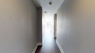 This two-floor high-rise residence offers a striking blend of contemporary design and urban living. The main living area is anchored by expansive floor-to-ceiling windows that frame breathtaking panoramic city skyline views, flooding the space with abundant natural light and casting dramatic shadows across the wide-plank hardwood flooring. A sleek staircase with modern metal railings connects both levels, adding an architectural focal point to the open layout.

The kitchen is thoughtfully designed with high-gloss white cabinetry, stone countertops, stainless steel appliances including a gas range, built-in microwave, and dishwasher, all arranged in an efficient galley-style configuration. Recessed and pendant lighting complete the polished look.

The hallways feature rich dark hardwood flooring, clean white trim, and recessed lighting, creating a refined transition between rooms. The bathroom is outfitted with a floating dark-toned vanity, a large wall mirror with overhead lighting, and an enclosed glass shower with a neutral tile surround.

A dedicated laundry area houses a full-sized washer and dryer, adding functional convenience. Upper-floor wraparound glazing offers an immersive elevated perspective of the surrounding cityscape. Wall-mounted sconce lighting in the living areas adds a warm, curated ambiance throughout.