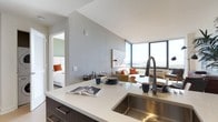 This thoughtfully designed single-floor residence showcases a seamless open-concept layout that integrates the kitchen, living, and dining areas into one cohesive and functional space. The contemporary kitchen features dark cabinetry paired with a crisp white countertop, a deep undermount sink, and a high-arc pull-down faucet — offering both style and practicality. The kitchen island serves as a natural gathering point and transitions effortlessly into the living and dining areas. The living space is anchored by a comfortable sofa accented with warm orange throw pillows, complemented by a curated selection of furniture and wall art that adds character to the neutral palette. The standout feature of the space is the expansive floor-to-ceiling corner windows that flood the interior with abundant natural light while framing striking panoramic views of the surrounding skyline and waterfront. A bedroom is visible through an open doorway, finished with a clean, modern aesthetic consistent with the rest of the residence. The overall ambiance is bright, airy, and sophisticated, combining clean architectural lines with warm accent tones to create a welcoming yet refined environment.