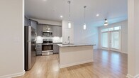 Located at 50 Fenway Place in Concord, North Carolina, this single-floor residence offers a thoughtfully designed open-concept layout with modern finishes throughout. The kitchen serves as the centerpiece of the living space, showcasing grey shaker-style cabinetry, granite countertops, a large central island with an undermount sink, and a full suite of stainless steel appliances including a refrigerator, range, microwave, and dishwasher. Stylish pendant lighting illuminates the island, adding a contemporary touch. Light hardwood-style flooring extends through the main living areas, creating a warm and cohesive aesthetic. The living area is bright and airy, featuring French doors with transom windows that invite natural light and provide outdoor access. The bedrooms offer plush carpeting and ceiling fans, with windows that bring in additional natural light. The bathroom continues the home's refined design language, featuring granite countertops, grey cabinetry with chrome hardware, an undermount sink, and a large mirror. A dedicated laundry area is also present within the unit. The overall ambiance is clean, modern, and move-in ready, with neutral tones and quality materials throughout creating a welcoming and functional living environment.