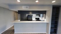 This single-story space showcases a thoughtfully designed kitchen that serves as a central feature of the layout. The kitchen is defined by a bold contrast of dark, charcoal-toned cabinetry paired with crisp white countertops, creating a contemporary aesthetic. A spacious peninsula counter extends outward, offering additional prep space and serving as a natural divider between the kitchen and the adjoining living area, reinforcing the open-concept design.

Black appliances, including a full-size refrigerator and an electric range with overhead ventilation, are seamlessly integrated into the cabinetry. Upper cabinets in matching dark tones line the walls, providing ample storage. Recessed lighting in the textured ceiling ensures the kitchen is well-lit, while a centrally located HVAC vent supports climate control throughout the space.

The flooring features durable wood-look planks that run throughout, adding warmth to the otherwise cool color palette. Light gray walls complement the cabinetry tones, contributing to a cohesive and modern interior. A doorway visible in the background hints at additional rooms, suggesting a functional and connected floor plan overall.