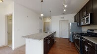 This single-floor residence offers a thoughtfully designed open-concept layout that seamlessly connects the kitchen and living areas. The kitchen serves as the centerpiece of the space, showcasing a large central island with granite countertops, an integrated sink, and pendant lighting overhead. Dark espresso cabinetry extends to the ceiling, providing generous storage, while a full suite of stainless steel appliances — including a refrigerator, gas range, built-in microwave, and dishwasher — delivers a modern, functional aesthetic. Warm hardwood flooring runs throughout the kitchen and dining area, transitioning to plush neutral carpeting in the spacious living room. The living area benefits from natural light streaming through a large double window and is equipped with a ceiling fan for year-round comfort. Track lighting complements the kitchen and entryway areas, adding both practicality and style. The bathroom features full white tile surrounding a soaking tub and shower combination, a dark-toned vanity with a white countertop, and clean tile flooring. The overall color palette — crisp white walls, dark cabinetry, and warm flooring tones — creates a balanced and inviting atmosphere throughout the home.