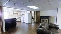 This single-floor space offers a practical and functional open-concept layout that seamlessly connects the living area and kitchen. The living space is anchored by a dark leather sofa positioned near a classic fireplace with a dark mantle and decorative surround, providing a focal point to the room. The kitchen is efficiently designed with light wood cabinetry, a white refrigerator, a stove with microwave overhead, and a countertop that doubles as a breakfast bar with bar stool seating — ideal for casual dining. A drop ceiling with recessed fluorescent lighting illuminates the space evenly throughout. Dark hardwood-style flooring runs continuously across the open floor plan, adding warmth and a cohesive visual flow. A separate room accessible through a solid wood door is visible toward the back of the space, offering a private area suitable for an office or additional use, as evidenced by the desk chair visible within. The overall layout is straightforward and versatile, making efficient use of the available footprint while maintaining a clear division between the social living and kitchen zones and the more private rear area.