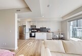 Located at 8450 106A Avenue NW in Edmonton, Alberta, this thoughtfully designed one-bedroom, one-bathroom unit makes excellent use of its compact footprint across a single floor. The open-concept layout flows seamlessly from the entry hallway — lined with a warm wood storage credenza — through a sleek, modern kitchen and into the living area beyond.

The kitchen is outfitted with white flat-panel cabinetry extending to the ceiling, a grey tile backsplash, a stainless undermount sink, and integrated appliances including a dishwasher. A bar-height table with stools is tucked beside a large sliding window, providing a bright dining nook with urban views of the Edmonton skyline.

The living area benefits from generous natural light through expansive windows, with light wood-look flooring running throughout the space, enhancing the airy, minimalist aesthetic. The spacious primary bedroom features a closet and is partially separated from the kitchen area by a structural column, offering a sense of privacy within the open plan.

The bathroom is finished to a high standard, featuring large-format grey wall tiles in the tub/shower surround, a floating white vanity with a bold matte black vessel sink, grey quartz countertop, built-in open shelving, and stone-look floor tiles. Black hardware accents are consistent throughout, lending a cohesive, contemporary character to the entire unit.