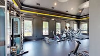 This fitness center is a thoughtfully designed workout facility that combines a bold aesthetic with a comprehensive range of exercise equipment. The space features a striking vaulted ceiling accented by a vivid yellow stripe that runs along the crown molding, complementing the deep charcoal-gray walls and dark tray ceiling details. Recessed lighting and a statement pendant fixture illuminate the room, while multiple large, white-trimmed windows line the walls, flooding the space with natural light and offering views of surrounding greenery.

The durable rubber flooring spans the entire room, providing a safe and practical surface for all types of training. Along one side, a row of Matrix treadmills is neatly arranged facing the windows, ideal for cardio workouts with a view. Spin bikes and elliptical machines offer additional cardio options, while a cable resistance machine and a full dumbbell rack stocked with a range of free weights support strength training. A power rack is positioned along the wall for barbell exercises.

A motivational "Stronger Than Yesterday" mural adds an energizing touch to one wall, reinforcing the facility's focus on progress and performance. The overall ambiance is modern, focused, and professional, making this fitness center a well-rounded destination for a complete workout experience.