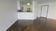 This single-floor residence showcases a thoughtfully designed open-concept layout that seamlessly connects the living area to the kitchen. Rich, dark hardwood flooring runs throughout the main living space, providing a warm and polished aesthetic that contrasts beautifully with the bright white walls.

The kitchen is defined by a raised peninsula that serves as both a functional workspace and a visual divider between the cooking and living areas. White cabinetry extends to the upper portion of the kitchen walls, offering generous storage, while dark stone countertops add a sophisticated contrast. A decorative tile backsplash introduces subtle texture and pattern to the space. Stainless steel appliances, including a refrigerator and microwave, contribute a modern, streamlined look. A flush-mount ceiling light provides focused illumination over the kitchen area.

Adjacent to the main living space, a doorway leads to an additional room, suggesting a versatile floor plan suited to various living arrangements. The overall ambiance is clean, contemporary, and move-in ready, with a neutral palette that allows for a wide range of interior design preferences.