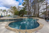 Located at 8104 Webb Rd in Riverdale, Georgia, this outdoor amenity area centers around a generously sized freeform swimming pool with a distinctive curvilinear design. The pool's shape incorporates a unique island feature with mature ornamental trees, adding a resort-like character to the space. Clear, blue water and multiple stainless steel entry points make the pool both visually inviting and accessible.

Surrounding the pool is a spacious paved sun deck finished in light-toned stonework, furnished with blue lounge chairs positioned to take full advantage of the open sky. Outdoor dining seating with a market umbrella is also available near the pool house, providing a comfortable spot for relaxation and socializing.

The pool house itself is a charming brick structure featuring an arched window, white trim, decorative railings, and a staircase entry — lending a classic, well-appointed aesthetic to the area. Manicured landscaping with mulched garden beds, trimmed hedgerows, and towering pine trees frames the entire space, offering a sense of natural enclosure and privacy.

The grounds are enclosed by a black iron fence, and views of the surrounding residential buildings are visible through the mature tree canopy, giving the area a well-integrated community feel. Overall, this pool area delivers an attractive and functional outdoor retreat.