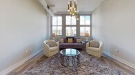 Located in the heart of downtown Dayton, Ohio, this stylish single-floor unit blends modern design with urban charm. The open-concept living area is a standout, featuring large multi-pane windows that frame sweeping city skyline views and flood the space with natural light. Exposed ductwork along the ceiling nods to the building's industrial roots, while warm hardwood flooring runs throughout, tying the space together cohesively. A striking cluster of gold pendant lights anchors the living area, complementing the contemporary furnishings below.

The kitchen is thoughtfully designed with flat-front cabinetry in a warm wood tone, sleek white countertops, a herringbone tile backsplash, and stainless steel appliances including a range, microwave, and refrigerator. Gold-toned fixtures add an elevated touch. A breakfast bar with seating provides a casual dining option and connects seamlessly to the main living space.

The bedroom is calm and understated, with light wood-look flooring, neutral walls, and a structural concrete column that reinforces the building's historic character. The bathroom is finished to a high standard, featuring large-format marble-look floor and wall tiles, a backlit vanity mirror, a modern single-sink vanity with gold accents, and a glass-enclosed tub-shower combination.
