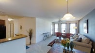 This single-floor residence offers a well-designed, open-concept layout that promotes a sense of spaciousness and ease of living. The living area is generously proportioned, featuring plush carpeted flooring and clean, neutral-toned walls that create a calm and inviting atmosphere. A ceiling fan with a light fixture is centrally positioned, complementing the stylish pendant light that anchors the dining area nearby.

Natural light flows abundantly through multiple large windows dressed with patterned curtains, brightening the interior and offering views to the outside. A glass door provides additional light and direct access to an outdoor area, enhancing the airy feel of the space.

The living room is designed for both comfort and functionality, with ample room for seating arrangements. A partial wall with a raised granite-topped counter defines the boundary between the kitchen and the living area, creating a casual breakfast bar while maintaining the open sightlines throughout.

The kitchen area, partially visible, features warm-toned countertops and a decorative arrangement that adds character to the space. Throughout the home, the consistent use of neutral colors, natural light, and thoughtful design elements come together to create a cohesive, comfortable, and welcoming living environment.