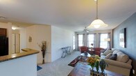 This single-floor residence offers a well-designed, open-concept layout that promotes a sense of spaciousness and ease of living. The living area is generously proportioned, featuring plush carpeted flooring and clean, neutral-toned walls that create a calm and inviting atmosphere. A ceiling fan with a light fixture is centrally positioned, complementing the stylish pendant light that anchors the dining area nearby.

Natural light flows abundantly through multiple large windows dressed with patterned curtains, brightening the interior and offering views to the outside. A glass door provides additional light and direct access to an outdoor area, enhancing the airy feel of the space.

The living room is designed for both comfort and functionality, with ample room for seating arrangements. A partial wall with a raised granite-topped counter defines the boundary between the kitchen and the living area, creating a casual breakfast bar while maintaining the open sightlines throughout.

The kitchen area, partially visible, features warm-toned countertops and a decorative arrangement that adds character to the space. Throughout the home, the consistent use of neutral colors, natural light, and thoughtful design elements come together to create a cohesive, comfortable, and welcoming living environment.