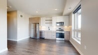 This single-floor unit showcases a thoughtfully designed, modern interior with a cohesive neutral palette throughout. The kitchen serves as a centerpiece, featuring flat-front cabinetry in a warm two-tone finish — white uppers paired with natural wood-toned lowers — complemented by a white subway tile backsplash, open shelving, and a full suite of stainless steel appliances including a gas range, over-the-range microwave, dishwasher, and refrigerator. A matte black faucet and undermount sink add a contemporary contrast. Rich, dark-toned hardwood-style flooring runs through the kitchen and living areas, creating a warm, grounded feel.

The living area opens to a private balcony through a large sliding glass door, with an adjacent window framing outdoor views, allowing ample natural light to fill the space. The bedroom features plush carpeting, a ceiling fan with light fixture, and generous natural light from a large window. A walk-in closet provides practical storage with built-in rod and shelving. The bathroom includes warm wood-toned cabinetry for added storage, hardwood-style flooring, and clean, minimalist finishes throughout.