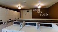 This communal laundry facility is designed with functionality and convenience in mind. The space is outfitted with a row of commercial-grade Speed Queen washers along the front, paired with a bank of matching dryers along the side wall, providing ample capacity to handle laundry needs efficiently. A long, continuous folding counter runs along the back wall, offering generous surface space for sorting and folding clothes. The room's interior features a bold, dark brown accent wall adorned with the text "WASH DRY FOLD," reinforcing the room's purpose while adding a touch of intentional design. Warm overhead flush-mount light fixtures illuminate the space, creating a bright and functional environment. The textured ceiling and neutral flooring contribute to the practical aesthetic of the room. A small ventilation panel is visible on the back wall, ensuring proper airflow throughout the facility. Overall, the space is clean, organized, and purpose-built to serve as a reliable shared laundry room, with all essential equipment and workspace thoughtfully arranged for user ease and efficiency.