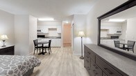 Located at 555 Executive Dr NW in Huntsville, Alabama, this single-floor studio unit offers a thoughtfully designed open-concept layout that efficiently combines living, sleeping, and dining areas into one cohesive space. Light wood-look flooring runs throughout, lending a clean and modern aesthetic that ties the entire unit together.

The sleeping area is comfortably sized to accommodate a full bed with matching nightstands and bedside lamps, while a window framing lush outdoor greenery fills the space with natural light. A wall-mounted HVAC unit ensures year-round climate comfort. Adjacent to the sleeping area, a sitting chair provides a dedicated relaxation spot, and a compact dining table with chairs offers a functional space for meals.

The kitchen is well-equipped with white cabinetry, a double-basin sink, a full range with electric stovetop, over-the-range microwave, and a refrigerator — providing all the essentials for everyday cooking. A dresser with mirror rounds out the living space, offering practical storage.

The bathroom features a single-sink vanity with white cabinetry, a large mirror with overhead lighting, and a tiled shower-tub combination, delivering a clean and functional design. The overall ambiance of the unit is bright, neutral, and move-in ready.