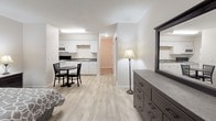 Located at 555 Executive Dr NW in Huntsville, Alabama, this single-floor studio unit offers a thoughtfully designed open-concept layout that efficiently combines living, sleeping, and dining areas into one cohesive space. Light wood-look flooring runs throughout, lending a clean and modern aesthetic that ties the entire unit together.

The sleeping area is comfortably sized to accommodate a full bed with matching nightstands and bedside lamps, while a window framing lush outdoor greenery fills the space with natural light. A wall-mounted HVAC unit ensures year-round climate comfort. Adjacent to the sleeping area, a sitting chair provides a dedicated relaxation spot, and a compact dining table with chairs offers a functional space for meals.

The kitchen is well-equipped with white cabinetry, a double-basin sink, a full range with electric stovetop, over-the-range microwave, and a refrigerator — providing all the essentials for everyday cooking. A dresser with mirror rounds out the living space, offering practical storage.

The bathroom features a single-sink vanity with white cabinetry, a large mirror with overhead lighting, and a tiled shower-tub combination, delivering a clean and functional design. The overall ambiance of the unit is bright, neutral, and move-in ready.
