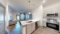 Located in Spring, TX, this well-appointed 1-bedroom, 1-bathroom residence at Casella offers a thoughtfully designed open-concept layout on a single floor. The kitchen serves as the heart of the home, featuring sleek gray shaker cabinetry, white quartz countertops, a subway tile backsplash, and a full suite of stainless steel appliances including a French door refrigerator, range, and built-in microwave. A central island with an undermount sink and seating for multiple bar stools makes it ideal for both cooking and casual dining. Pendant lighting overhead adds a modern, polished touch. The kitchen flows seamlessly into the living area, which benefits from large windows that invite abundant natural light and provide outdoor views. Wood-style plank flooring extends throughout the main living areas, lending a warm, cohesive aesthetic. The generously sized bedroom features plush carpet, multiple windows, and ample room for a full furniture arrangement. The bathroom offers a quartz vanity, an undermount sink, a large framed mirror, and a glass-enclosed tiled shower. A spacious walk-in closet with dual hanging rods and built-in shelving provides excellent storage. A dedicated in-unit laundry room with side-by-side washer and dryer and overhead shelving adds everyday convenience.