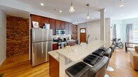 Located at 14 Homer St in Brookline, MA, this two-bedroom, two-bathroom home spans two floors, offering a well-organized and functional layout. The main level features warm hardwood flooring throughout and includes a combined kitchen and living area anchored by a granite-topped island with bar seating. The kitchen is equipped with stainless steel appliances, rich wood cabinetry, a gas range, and is accented by an exposed brick wall that adds character to the space. Adjacent to the kitchen, the dining room is filled with natural light from multiple windows dressed with soft grey curtains and is centered by a classic chandelier overhead. The primary bedroom on the main level continues the hardwood flooring and features a closet and recessed lighting. Both bathrooms are finished with marble-style tile and include shower enclosures. The lower floor provides the second bedroom, a generously proportioned room with neutral carpet, ample space, and direct access to additional areas. A hallway and two staircases connect the floors efficiently. Overall, the home presents a blend of classic architectural details, including exposed brick, alongside modern finishes, creating a comfortable and inviting living environment.