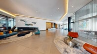 This contemporary lobby is a masterclass in modern design, offering an expansive, open floor plan finished with large-format neutral floor tiles that amplify the sense of space. The centerpiece is a sleek reception desk dramatically backdropped by a floor-to-ceiling geometric gold-tiled accent wall, warmly illuminated to draw the eye immediately upon entry. Ambient cove lighting runs along the ceiling perimeter, complementing recessed downlights to create a layered, inviting atmosphere throughout.

A dedicated lounge area anchors one side of the lobby, featuring a wide, linear fireplace set beneath an elegant marble-finish surround, accented by sculptural wall art in vibrant jewel tones. Circular pendant lighting adds a contemporary focal point overhead. Nearby, velvet seating in bold rust and teal tones is arranged across curated area rugs, offering comfortable conversation areas. Sheer floor-to-ceiling curtains filter abundant natural light from expansive glazed facades, while semi-transparent chain curtain partitions define a connected co-working or lounge zone without sacrificing openness.

A private dining nook features a live-edge wood communal table surrounded by upholstered rust-toned chairs, bathed in natural light from glass walls overlooking an exterior drop-off area. Large-format stone wall cladding and dark structural columns add depth and textural contrast throughout, completing this polished, thoughtfully curated environment.