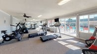 This fitness center is a dedicated, single-floor exercise facility designed to support a well-rounded workout routine. The space is bright and airy, benefiting from an abundance of natural light that streams in through large sliding glass doors and wide windows lining the walls, offering views of the adjacent outdoor pool and surrounding landscape. The interior is finished with light grey walls, crown molding, and a textured ceiling, giving the room a clean and well-maintained appearance. Ceiling fans are positioned throughout the space to promote airflow and comfort during workouts. The floor is covered with durable carpet and rubberized equipment mats, providing traction and cushioning underfoot. A full-length wall mirror on one side visually expands the room and assists users in monitoring their form. The cardio equipment selection includes multiple treadmills, a stationary recumbent bike, an upright exercise bike, and an air resistance bike, accommodating a variety of fitness preferences. A dumbbell rack with a range of free weights is available for strength training. A wall-mounted flat-screen television is positioned for viewing during cardio sessions. Safety equipment, including a fire extinguisher, is visibly mounted on the wall. The facility's direct access to the outdoor pool area further enhances its appeal as a comprehensive wellness amenity.