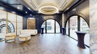 Located at 28 N Saginaw St in the heart of downtown Pontiac, Michigan, this sophisticated single-floor event space delivers a striking blend of classic grandeur and contemporary elegance. The interior is defined by deep navy blue wall paneling accented with polished gold trim, creating a rich and refined atmosphere throughout. Herringbone-patterned hardwood-style flooring runs the length of the space, adding texture and warmth underfoot.

Dramatic arched floor-to-ceiling windows line the street-facing walls, flooding the interior with natural light and offering views of the vibrant downtown streetscape. Between the arches, columns finished in veined marble add architectural depth and contrast beautifully against the dark paneling. Multiple oversized circular crystal chandeliers hang from the coffered ceilings, providing a glamorous focal point and warm ambient lighting.

The space features several distinct lounge and gathering areas furnished with cream and gold velvet seating, marble-topped accent tables, and tall navy cocktail tables positioned near the windows. A gallery wall of large gold-framed oil paintings depicting elegant social scenes adds a curated, artistic character to one section of the space. A service counter is also present, supporting catering and event needs. This thoughtfully designed venue is well-suited for receptions, private events, and upscale social functions.