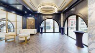 Located at 28 N Saginaw St in the heart of downtown Pontiac, Michigan, this sophisticated single-floor event space delivers a striking blend of classic grandeur and contemporary elegance. The interior is defined by deep navy blue wall paneling accented with polished gold trim, creating a rich and refined atmosphere throughout. Herringbone-patterned hardwood-style flooring runs the length of the space, adding texture and warmth underfoot.

Dramatic arched floor-to-ceiling windows line the street-facing walls, flooding the interior with natural light and offering views of the vibrant downtown streetscape. Between the arches, columns finished in veined marble add architectural depth and contrast beautifully against the dark paneling. Multiple oversized circular crystal chandeliers hang from the coffered ceilings, providing a glamorous focal point and warm ambient lighting.

The space features several distinct lounge and gathering areas furnished with cream and gold velvet seating, marble-topped accent tables, and tall navy cocktail tables positioned near the windows. A gallery wall of large gold-framed oil paintings depicting elegant social scenes adds a curated, artistic character to one section of the space. A service counter is also present, supporting catering and event needs. This thoughtfully designed venue is well-suited for receptions, private events, and upscale social functions.