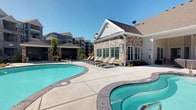 This outdoor amenity area is designed to deliver a resort-like experience for residents. At its center is a generously sized, freeform swimming pool with vivid turquoise water, complemented by a separate spa or hot tub that shares the same curvilinear design language. The surrounding concrete sundeck provides ample space for relaxation, lined with a row of chaise lounge chairs ideal for sunbathing on clear days.

The adjacent single-story clubhouse serves as an architectural anchor for the space, showcasing a refined combination of natural stone veneer and white board-and-batten siding beneath a steeply pitched, shingle roof. Large windows along the clubhouse facade allow natural light to filter inside while maintaining a visual connection to the pool area. A covered pergola or shade structure at the pool's edge offers a respite from direct sun.

The backdrop features multi-story residential buildings with a modern farmhouse aesthetic, consistent in their gray siding and pitched rooflines. Thoughtful landscaping with young trees and greenery softens the hardscape, while the expansive open sky above enhances the bright, airy atmosphere of the entire outdoor space.