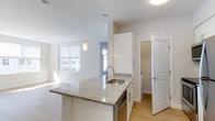 This one-bedroom, one-bathroom residence with a den is designed with an open, efficient layout spread across a single floor. The kitchen serves as the focal point of the living space, anchoring the open-concept floor plan with a generously sized central island topped with light-veined granite countertops. A stainless steel undermount sink and modern arc faucet are integrated into the island, which also accommodates a dishwasher beneath. White shaker-style cabinetry extends to the ceiling, maximizing storage, while pendant lights with warm-toned shades hang above the island to provide focused task lighting. Light hardwood flooring runs throughout, contributing to a cohesive and airy aesthetic. The kitchen flows naturally into the living area, which receives abundant natural light through large windows fitted with horizontal blinds. The open sightlines between the kitchen and living space create a sense of generous square footage. A paneled interior door leads to an additional room, likely the den, offering a versatile space suitable for a home office or guest area. The overall palette of white walls, light flooring, and neutral granite creates a clean, modern ambiance throughout.