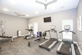 This dedicated fitness center is thoughtfully designed to support a full range of workout routines. The space features durable rubber flooring throughout, providing both safety and functionality for exercise activities. Neutral gray walls and recessed lighting create a clean, focused atmosphere, while a ceiling fan ensures comfortable airflow during workouts.

The room is outfitted with a strong selection of professional-grade True brand cardio equipment, including a treadmill, elliptical, and rowing machine, catering to a variety of aerobic training preferences. A multi-function strength rack with an adjustable bench and a full dumbbell rack with a wide range of weights address all free weight and resistance training needs. A wall-mounted flat-screen television adds entertainment during workouts, and a beverage refrigerator stocked with water bottles keeps hydration convenient.

Full-length mirrors are positioned along the walls, allowing users to monitor form during exercise. Large interior windows connect the fitness room to an adjacent lounge or common area, creating an open, airy feel. A separate hallway with additional doors suggests access to restroom or changing facilities nearby. The overall design is practical and modern, delivering a well-rounded fitness environment suitable for daily use.