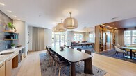 Located at 437 Cold Brook Dr in Sudbury, MA, this single-floor community amenity space is thoughtfully designed to offer a variety of social and recreational experiences. The open-concept layout connects several distinct zones while maintaining a cohesive, contemporary aesthetic throughout.

The central dining area is anchored by a large, elongated dark table surrounded by mixed seating and topped with a trio of oversized woven pendant lights suspended from brass fixtures — a striking architectural focal point. Light hardwood flooring runs throughout the main areas, complementing the warm neutral palette of the walls and furnishings.

Adjacent to the dining area is a well-appointed wet bar with light wood cabinetry, open shelving, and a textured tile backsplash — ideal for hosting and catering needs. A dedicated game room features a full-size billiards table beneath contemporary stacked pendant lighting, flanked by bar-height counter seating along the perimeter windows.

The lounge area transitions into a more relaxed setting, with a warm wood-paneled ceiling, built-in shelving with a mounted television, and an assortment of curved lounge chairs and modular seating in rich orange and neutral tones. Plush patterned carpeting defines the seating zones. Large windows throughout the space invite ample natural light, offering views of the surrounding outdoor areas and creating a bright, welcoming atmosphere.
