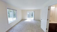 This two-story home features 2 bedrooms and 1.5 bathrooms, offering a practical and comfortable layout spread across two levels. The main living area is finished with neutral wall tones and plush carpeting, creating a warm and inviting atmosphere throughout. Large windows allow abundant natural light to fill the space, enhancing the open, airy feel of the interior. A sliding glass door provides direct access to the outdoors, seamlessly connecting the living area to the exterior and allowing views of the surrounding greenery. The clean, simple architectural lines and light color palette contribute to a bright and understated aesthetic. The half bathroom on the main level adds everyday convenience, while the full bathroom serves the upper floor. The two bedrooms offer private retreats, suitable for rest or use as a home office. Overall, the home presents a straightforward, functional design with well-maintained finishes and a layout that maximizes usability across both floors, making it a practical living space with a calm and comfortable ambiance throughout.