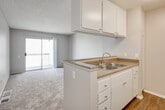 Located at 11902 Central Ave in Chino, California, this single-floor residence offers a thoughtfully arranged open-concept layout that seamlessly connects the living and kitchen areas. The kitchen is equipped with a peninsula-style counter featuring a double stainless steel sink, speckled granite-toned countertops, and white cabinetry with ample upper and lower storage. Wood-style flooring grounds the kitchen area, while built-in open shelving along the living room wall provides convenient display and storage space. The spacious living area is finished with plush wall-to-wall carpeting and a textured ceiling, and opens directly onto a private balcony through sliding glass doors, allowing natural light to fill the room. A well-sized bedroom continues the carpeted flooring and textured ceiling, featuring a window with blinds that brings in additional natural light. The bathroom is outfitted with a single-sink vanity topped with matching speckled countertops, white cabinetry, a large mirror, and a full shower-tub combination. Wood-style flooring in the bathroom and kitchen adds a cohesive design element throughout the unit. The neutral color palette and functional layout make this a comfortable and practical living space.