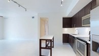 This 3-bedroom, 2-bathroom single-floor residence offers a clean, contemporary design with an open-concept layout that maximizes livability and flow. The kitchen is a standout feature, outfitted with rich dark cabinetry, stainless steel appliances, and light-toned countertops that create a striking contrast. A built-in breakfast bar with a matching dark wood base serves as a natural divider between the kitchen and the expansive living area, providing both a functional dining space and an architectural focal point. The living area is finished with large-format light tile flooring laid in a diagonal pattern, lending a sense of spaciousness and elegance throughout. Track lighting is installed across the ceiling, offering flexible and adjustable illumination suited to a variety of uses. The walls are finished in a neutral warm white, contributing to the bright and airy ambiance of the space. A hallway visible from the main living area leads to the private bedrooms and bathrooms, maintaining a clear and logical separation between living and sleeping spaces. Overall, the residence presents a well-appointed, modern aesthetic with thoughtful design details and a cohesive, light-filled atmosphere.