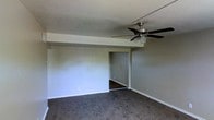 This single-floor space presents a straightforward and functional layout designed for comfortable everyday living. The main living area is finished with wall-to-wall carpet in a neutral gray-brown tone, providing a soft and practical underfoot surface. Smooth, light-colored walls create a clean and understated backdrop throughout the space, contributing to a bright and open feel. A ceiling fan with an integrated light fixture is centrally mounted overhead, offering both air circulation and ambient lighting to the room. Crisp white baseboards run along the perimeter of the walls, adding a clean finishing detail to the space. A distinct step-down or beam feature divides the ceiling plane, giving the room a subtle architectural element. An open doorway leads to an adjoining room, visible in the background with matching flooring and wall tones, suggesting a connected and cohesive interior flow. The overall ambiance of the space is practical and move-in ready, offering a neutral canvas that lends itself well to a variety of furnishing styles and personal touches. The single-floor layout ensures easy accessibility and convenient navigation throughout.