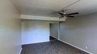 This single-floor space presents a straightforward and functional layout designed for comfortable everyday living. The main living area is finished with wall-to-wall carpet in a neutral gray-brown tone, providing a soft and practical underfoot surface. Smooth, light-colored walls create a clean and understated backdrop throughout the space, contributing to a bright and open feel. A ceiling fan with an integrated light fixture is centrally mounted overhead, offering both air circulation and ambient lighting to the room. Crisp white baseboards run along the perimeter of the walls, adding a clean finishing detail to the space. A distinct step-down or beam feature divides the ceiling plane, giving the room a subtle architectural element. An open doorway leads to an adjoining room, visible in the background with matching flooring and wall tones, suggesting a connected and cohesive interior flow. The overall ambiance of the space is practical and move-in ready, offering a neutral canvas that lends itself well to a variety of furnishing styles and personal touches. The single-floor layout ensures easy accessibility and convenient navigation throughout.