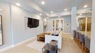 This single-floor space is thoughtfully designed with a contemporary aesthetic, offering a seamless flow between its main areas. The living room features soft, light gray walls, recessed lighting, and a tray ceiling that adds architectural depth. A wall-mounted flat-screen television is set into a dedicated niche, anchoring the seating area, which includes a neutral-toned sofa, a layered area rug, and coordinating side tables. Glass-paned French doors connect the living area to the rest of the space, maintaining an open and connected feel throughout.

The dining area is a standout with its raised tray ceiling, painted in a deeper shade of blue-gray that contrasts elegantly with the surrounding walls. A polished chrome lantern-style chandelier serves as a focal point above a round dining table surrounded by upholstered chairs. Large windows flood the room with natural light, while decorative circular mirrors and a lush fiddle-leaf fig tree add warmth and character.

The kitchenette is finished with granite countertops, a teal tile backsplash in a scalloped pattern, stainless steel appliances, and open floating shelves. Light-toned large-format tile flooring unifies all areas, contributing to the space's clean, modern, and welcoming ambiance.