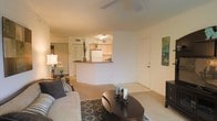 This single-floor residence is designed with an open-concept layout that seamlessly connects the living area to the kitchen. The living room is comfortably sized, featuring wall-to-wall carpeting that adds warmth underfoot, and is equipped with a ceiling fan overhead. A patterned area rug anchors the seating arrangement, which includes a plush sofa and a round dark-wood coffee table, creating a welcoming focal point.

The kitchen is accessible via a raised breakfast bar with a dark wood-toned countertop, offering a functional pass-through that maintains visual openness between the two spaces. White cabinetry and a white refrigerator are visible, giving the kitchen a clean, classic look. Recessed and overhead lighting ensures the kitchen remains well-lit.

The walls throughout are finished in a neutral, warm tone that complements the carpeted floors and contributes to a cohesive, understated ambiance. A hallway visible from the living area suggests additional rooms beyond the main living space. The overall design prioritizes practicality and an open feel, making the space functional and easy to navigate.